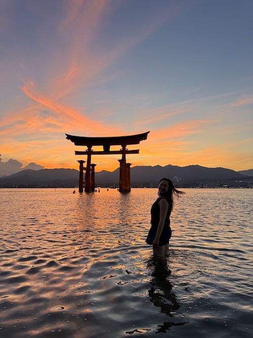 Torii on the water with sunset in the background and silhouette of a girl in the foreground.